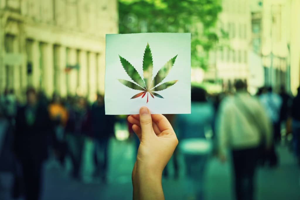 Hand holding a paper sheet with marijuana leaf symbol over a crowded street background. Cannabis legalization as medical drug. CBD healing social issue concept.