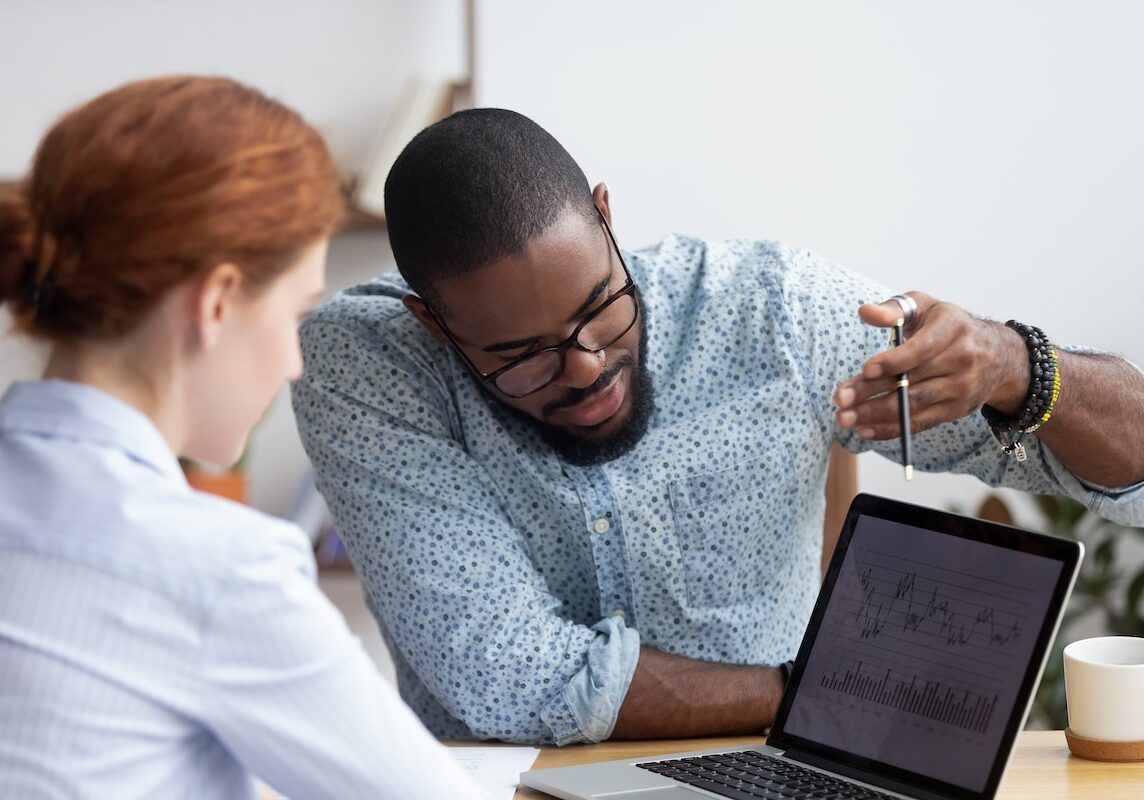 Diverse millennial colleagues working together analyzing diagram looking at computer screen. Black mentor helps female apprentice understand corporate program explaining interface showing on monitor