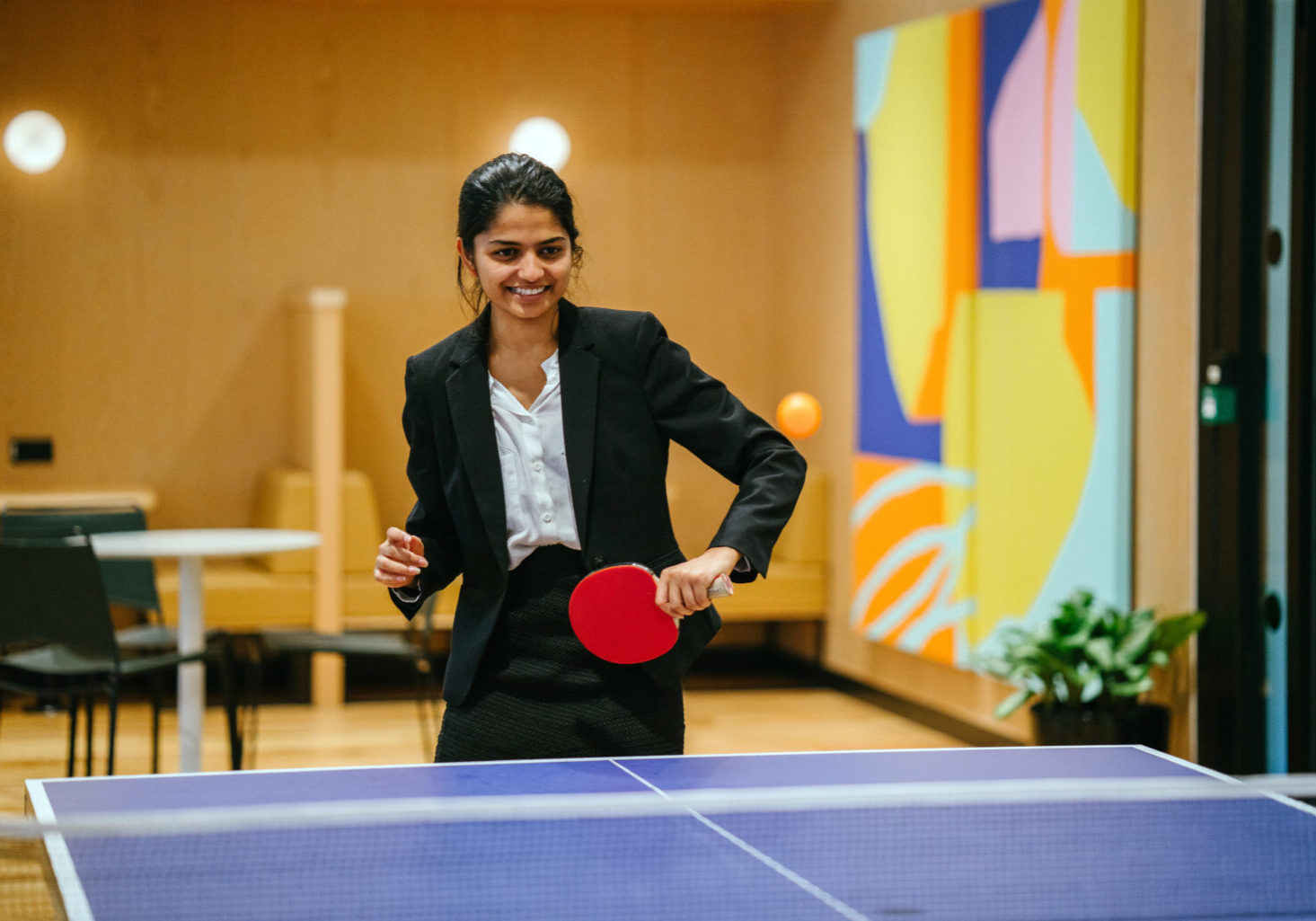 Portrait of a young and attractive Indian Asian woman in a suit playing table tennis with her colleague in the office during a break. She is having lots of fun and is smiling broadly.