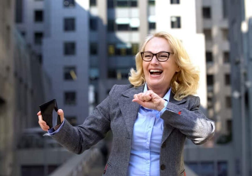 Blonde woman in a gray blazer laughing while holding a phone, with apartment buildings behind her