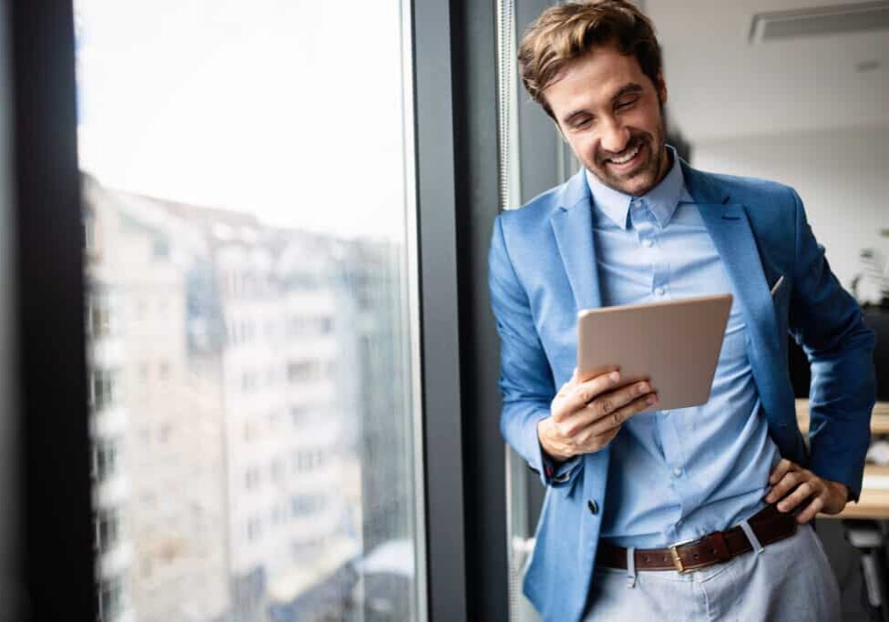 Man in a blue blazer smiling at a tablet while standing by an office window overlooking apartment buildings