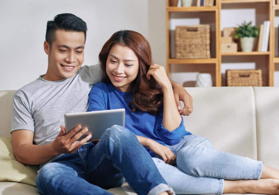 asian couple sitting on couch in apartment, smiling at tablet