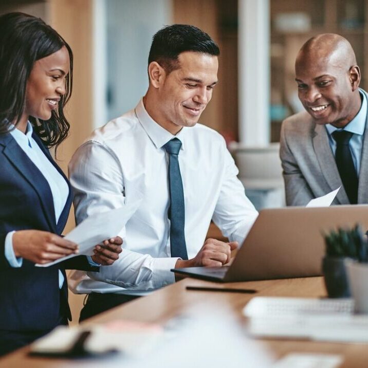 Smiling group of diverse businesspeople going over paperwork together and working on a laptop at a table in an office